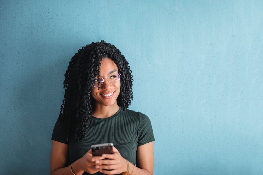 A cheerful young woman with curly hair uses her phone against a blue background.